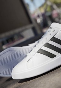 Weißer Sneaker mit schwarzen Streifen und weißen Schnürsenkeln auf grauer Oberfläche, ein Teil eines Schuhs zeigt die Sohle mit einem Fischgrätmuster.