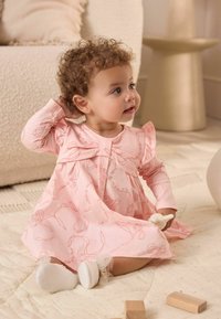 Abito rosa chiaro con maniche a volant e dettaglio a fiocco, realizzato in un tessuto morbido e caratterizzato da un delicato motivo a fiocco; il bambino è seduto su una superficie testurizzata.