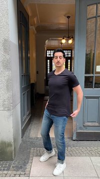 Man in black t-shirt and jeans stands holding his shirt, in front of a blue door with glass panels and a light fixture inside hallway.
