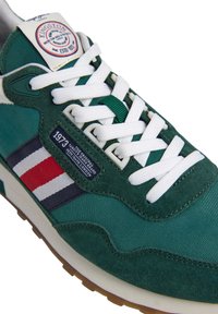 Baskets vertes avec lacets blancs, rayures latérales rouge, blanc et bleu, et étiquettes de marque "Kingston" et "1973" sur la languette et le panneau latéral.