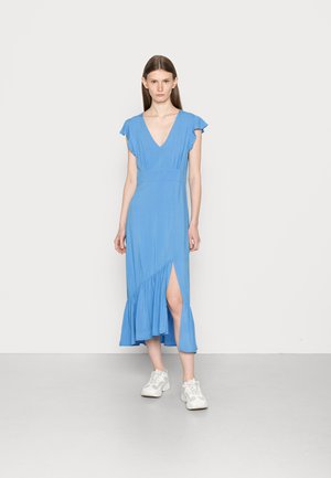 Vestido azul de comprimento até os joelhos com mangas em forma de braço, decote em V, pregas na cintura e uma barra com pregas. Combinado com ténis brancos.