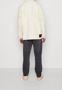 Chaqueta color crema con cuello abotonado, etiqueta de tela de contraste y jeans gris oscuro. Diseño ajustado con una textura suave.