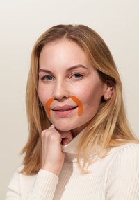 Modell med långt blont hår och en ribbad krämfärgad pulli, har orangea klisterlappar som liknar en mustasch på sitt ansikte.