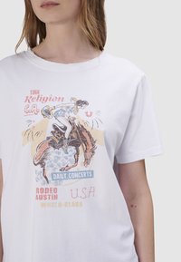 Wit katoenen T-shirt met een bedrukte rodeo-afbeelding van een ruiter op een paard, met pastelkleurige accenten en tekstdetails.