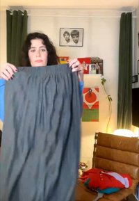 Kobieta z kręconymi włosami trzymająca szarą spódnicę w salonie z oprawionymi zdjęciami, zielonymi zasłonami i brązowym skórzanym fotelem.