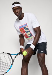 Tricou alb din bumbac cu un design grafic colorat. Șorturi negre cu logo-ul Nike. Ținând o minge de tenis și o rachetă, purtând o bandă de cap.