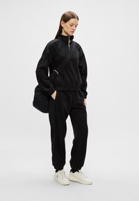 Femeie îmbrăcată într-un trening negru cu fermoar, cu mâinile în buzunare, adidași albi și geantă crossbody, stând pe un fond simplu, deschis la culoare.