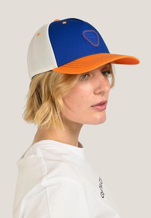 Personne blonde portant une casquette de baseball bleue et orange avec des côtés en maille blanche et un t-shirt blanc, regardant vers la droite.