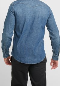 Mörkblå denimskjorta, långa ärmar, med knappmanschetter, en rundad nederkant och en yoke-detalj på baksidan. Textur av bomullstyg.