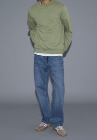Felpa verde con polsini e orlo a costine, indossata sopra una t-shirt bianca, abbinata a jeans blu a gamba dritta e scarpe da ginnastica grigie.