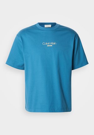 Niebieska bawełniana koszulka z krótkim rękawem i okrągłym dekoltem, z logo "Calvin Klein JEANS" w żółtym haftowanym napisie, umieszczonym na środku klatki piersiowej.