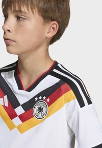 White soccer jersey with a V-neck, featuring black, red, and yellow diagonal stripes, and a round embroidered logo on the left chest.