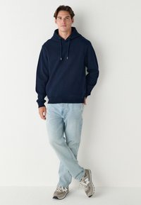 Laivastonsininen huppari teksturoidusta kankaasta, yhdistetty vaaleansinisiin farkkuihin ja harmaisiin lenkkareihin, joissa on valkoiset yksityiskohdat ja verkkokuviointi.