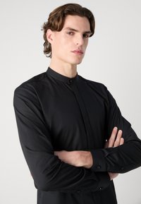 Crna majica dugih rukava s visokim ovratnikom, prednjim dugmadi i suptilnim sjajem, s glatkom teksturom i dizajnom koji se slim fit.