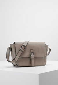 Taupe kožená křiž-crossbody taška s nastavitelným popruhem na přezku a detailní přezkou na přední straně, umístěná na bílé ploše a s neutrálním pozadím.