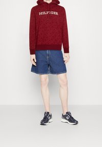 Sudadera roja con estampado y el texto "HILFIGER", combinada con shorts de mezclilla azules y zapatillas deportivas navy, que muestran un ajuste relajado y un estilo casual.