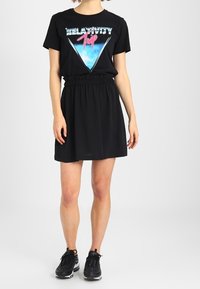 Abito t-shirt nero con un design grafico che presenta il testo "RELATIVITY", cintura elastica e maniche corte. Indossato con scarpe da ginnastica nere.