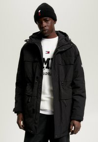 Parka negra aislante con capucha, con bolsillos frontales y cremallera, llevada sobre un suéter de punto blanco con texto de logo. Gorro negro en la cabeza.