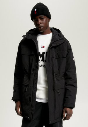 Parka negra aislante con capucha, con bolsillos frontales y cremallera, llevada sobre un suéter de punto blanco con texto de logo. Gorro negro en la cabeza.