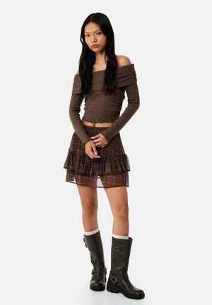 Jonge vrouw die een bruin gebreide off-shoulder top, een gelaagde bruine rokje met ruches en zwarte mid-kuitlaarzen met witte sokken draagt, staand tegen een witte achtergrond.