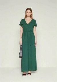 Maksikleita tumši zaļā krāsā ar V veida kakla izgriezumu, īsām plūstošām piedurknēm, savilktu jostasvietu ar saišu jostu un plūstošu svārku.