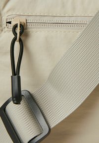 Beige Stofftasche mit einem Reißverschlussfach, strukturiertem Tragegurt und schwarzen Hardware-Details. Beinhaltet ein strapazierfähiges Elastikband für ein einfaches Öffnen.