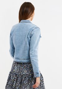 Chaqueta de mezclilla azul claro con mangas largas, diseño corto y cuello clásico. La parte posterior cuenta con una textura suave y no tiene tachuelas.
