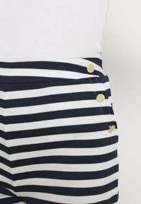 Námořnické a bílé pruhované bavlněné kalhoty s přiléhavým pasem, které mají tři zlaté knoflíky na levé straně a hladkou strukturu.