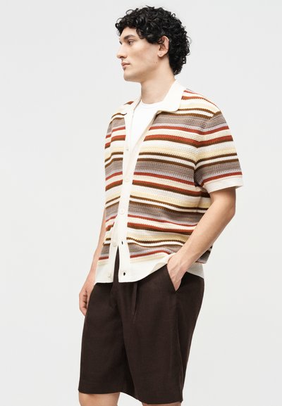 Άνδρας με σγουρά μαλλιά, που φοράει ριγωτό κοντομάνικο πλεκτό μπλουζάκι πάνω από λευκό μπλουζάκι και σκούρα καφέ σορτς μέχρι το γόνατο, στέκεται με τα χέρια στις τσέπες.