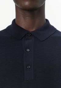 Polo de culoare albastru închis, cu tricot texturat, având o cămașă cu guler, trei butoane negre și o croială ajustată.