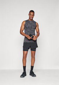 Fitted svart ermeløs topp med kamuflasjemønster, sammen med svart shorts. Modellen holder en svart vannflaske, iført sorte sokker og sneakers.