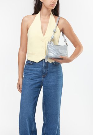Vrouw die een lichtgele mouwloze gilet en blauwe jeans draagt, met een zilveren gevlochten handtas met een geknoopte band over haar schouder.