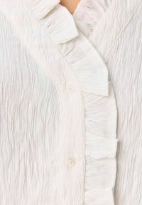 Blusa bianca in tessuto texturizzato con colletto a volant e pattina frontale, dotata di chiusura con un singolo bottone e sottili pince orizzontali.