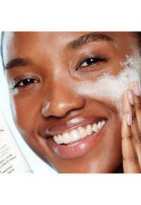 Close-up of a person smiling while applying a foamy facial cleanser. Smooth skin texture, light brown complexion, and short dark hair.