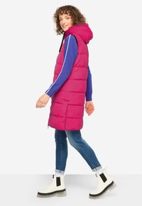 Fuchsia pufferväst med huva, med quiltat mönster och svart dragsko, bärs över en blå långärmad skjorta och kombineras med jeans och kängor.