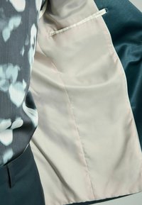 Blazer a medida con un exterior en azul petróleo oscuro y un forro de satén suave en beige claro. Presenta un sutil patrón floral en la tela exterior.