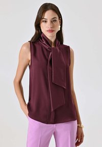 Blusa senza maniche color bordeaux con dettaglio decorativo di sciarpa annodata al collo. Tessuto liscio con vestibilità morbida, abbinata a pantaloni rosa chiaro.