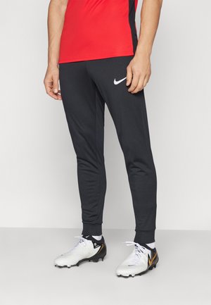 Schwarze Sporthose von Nike mit elastischen Bündchen, kombiniert mit weißen Fußballschuhen mit schwarzen und goldenen Akzenten. Rotes Oberteil ist teilweise sichtbar.