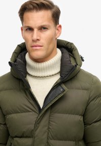 Chaqueta acolchada verde oliva con un alto cuello y cierre de cremallera, superpuesta sobre un suéter de punto acanalado color crema, que presenta acentos negros en la capucha.