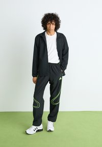 Sort atletisk jakke med limegrønne detaljer og elastiske bukser; båret med en hvid t-shirt og hvide sneakers med sorte detaljer.