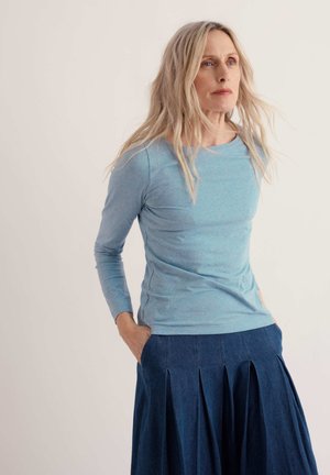 Femme aux longs cheveux blonds portant un haut bleu clair à manches longues et une jupe plissée bleue, debout, les mains dans les poches de la jupe, sur fond uni.