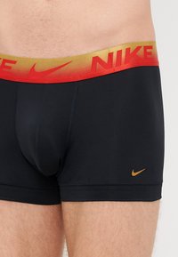 Boxeri negri cu un brâu elastic roșu și auriu care prezintă logo-ul "NIKE", logo-ul auriu Nike swoosh pe partea stângă, dintr-un material elastic.