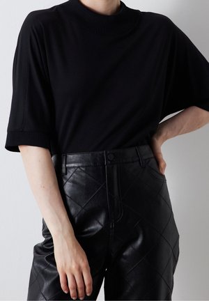 Persona vestida con un suéter negro de manga corta y pantalones de cuero acolchado negros de talle alto, con la mano en la cadera y relajada a un lado.