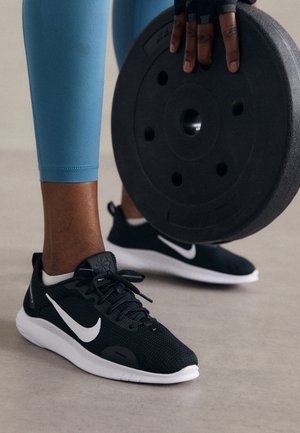 Osoba nosząca czarne sneakersy Nike i niebieskie legginsy podnosi dużą czarną tarczę do ćwiczeń w rękawiczce.