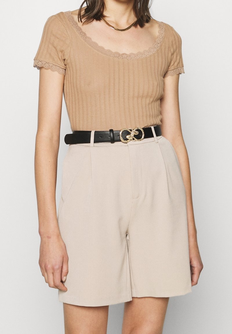 Femme portant un haut beige côtelé à manches courtes avec une bordure en dentelle, un short beige taille haute et une ceinture noire avec une boucle double anneau dorée.