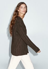 Donkerbruine kabelgebreide cardigan met een losse pasvorm, lange mouwen en een open voorkant. Gecombineerd met witte broek. Effen achtergrond.