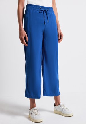 Niebieskie szerokie spodnie o skróconej długości, z elastycznym paskiem i sznurkiem, noszone z białymi sneakersami i białą bluzką.