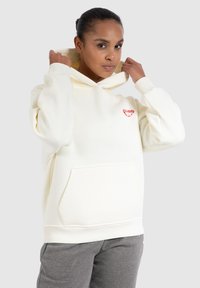 Felpa bianca realizzata in tessuto morbido, con un logo a forma di cuore rosso, tasca frontale e cappuccio con cordino. Accompagnata da pantaloni della tuta grigi.