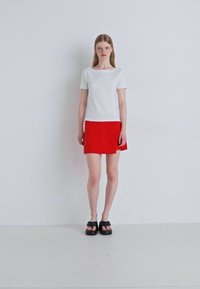 Bílý tričko s krátkým rukávem v kombinaci s červenou skater sukní. Modelka má na sobě černé platformové sandály. Minimalistický design s čistými liniemi.