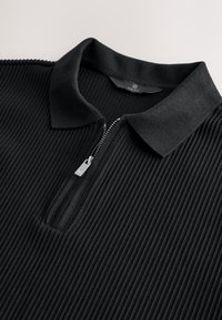 Schwarzes Poloshirt mit Reißverschluss am Kragen, das eine gerippte Textur und einen dezenten Glanz aufweist. Silberne Reißverschlussdetails verleihen dem Shirt Akzente.